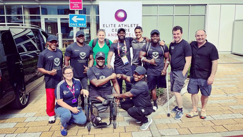 11 people posing for the camera and smiling outside the Elite Athlete Centre including Para athletes, coaches, practitioners and researchers